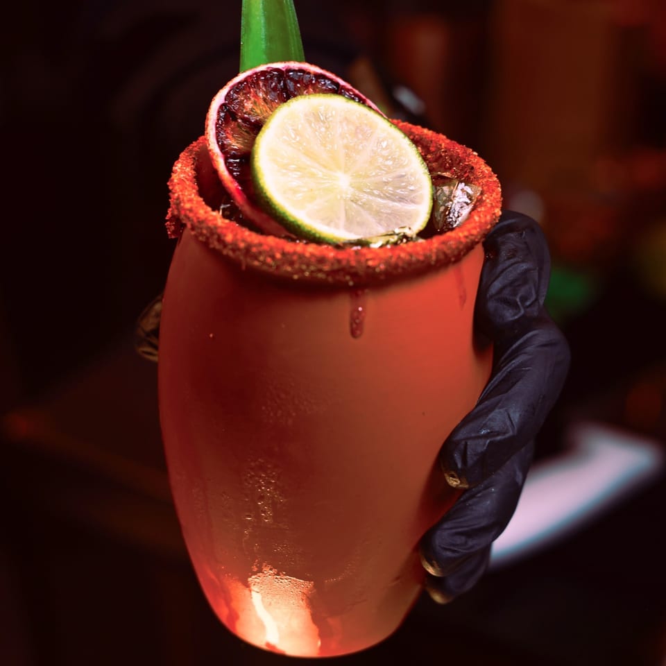 A hand in a black glove holds a Xochitl Cocina & Tequila cocktail with lime and orange slices.